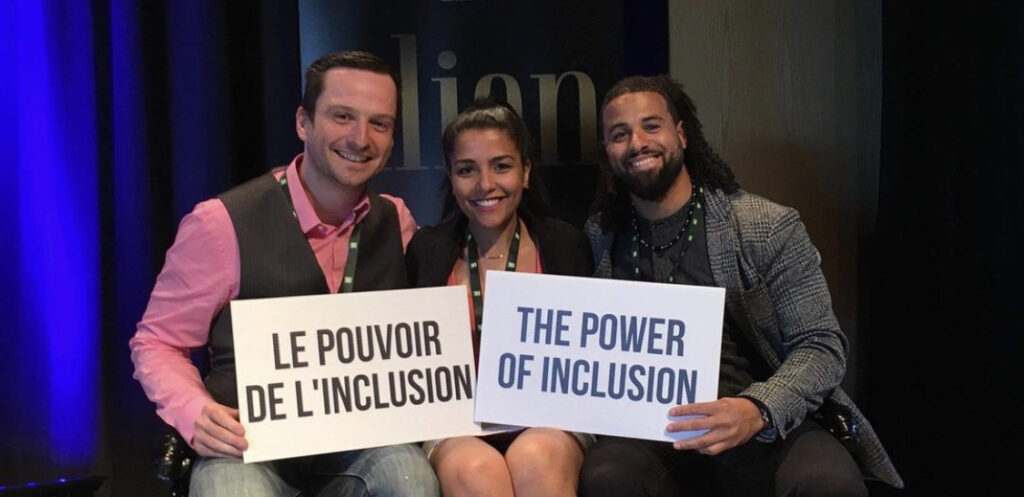 3 people, two male with female in centre sit close together smiling, holding two signs that say "The power of Inclusion" in both English and French